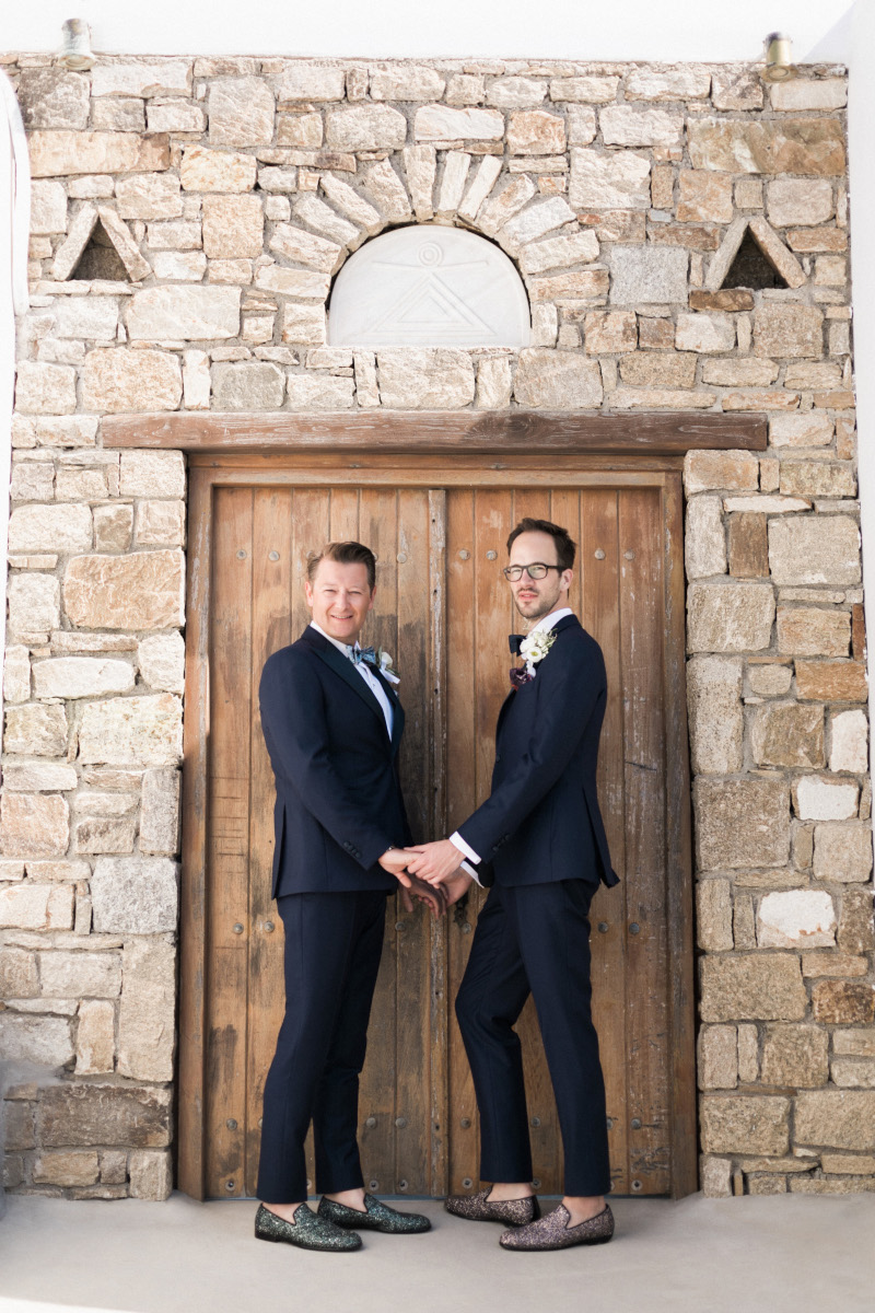 A gay couple of two men holding hands in front of a doorframe on their same sex Mykonos beach wedding 