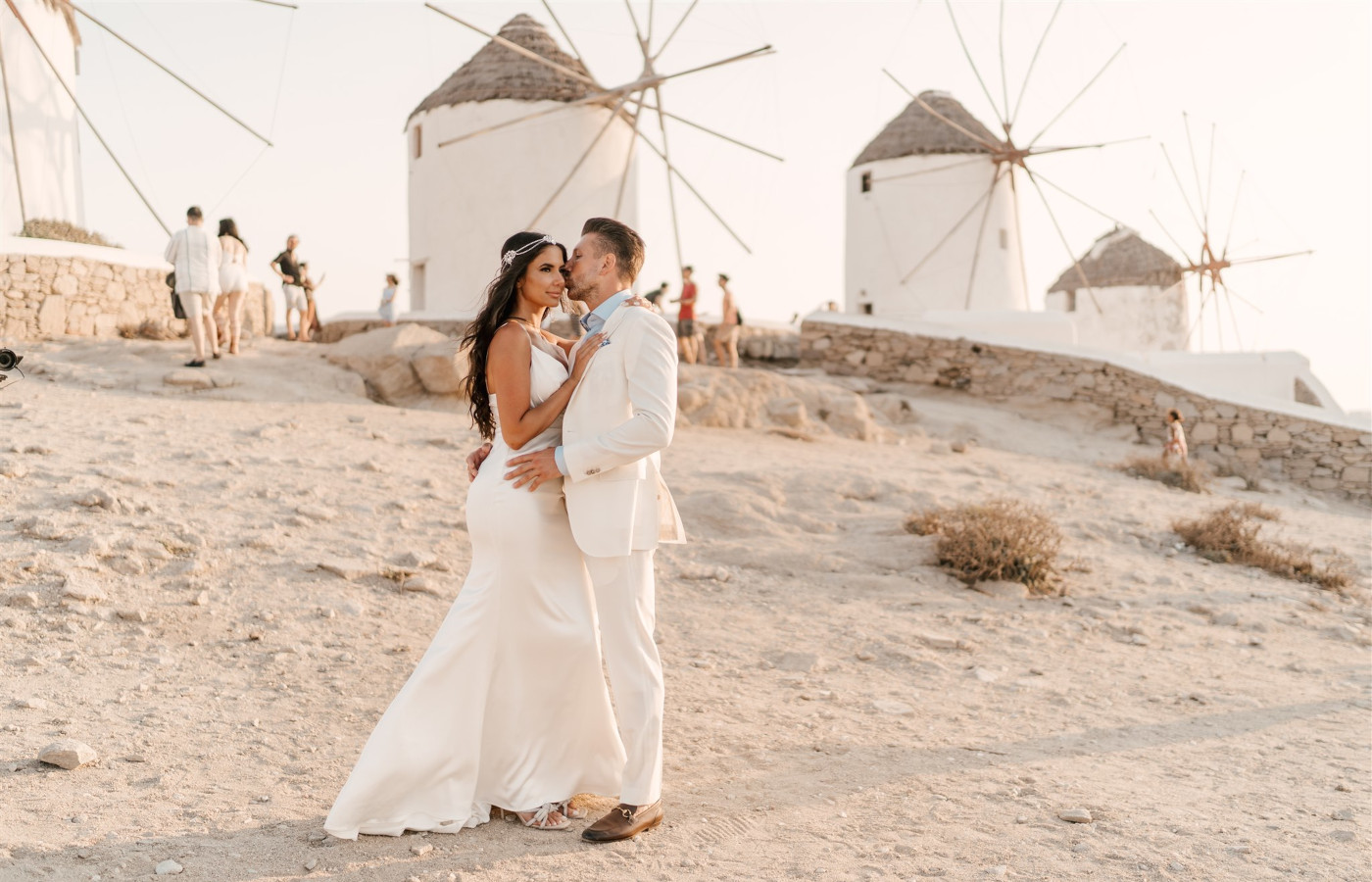 A groom in a white suit kissing his beautiful bride on the cheek, in front of Mykonos windmills, in the dream destination wedding in Mykonos Greece. 